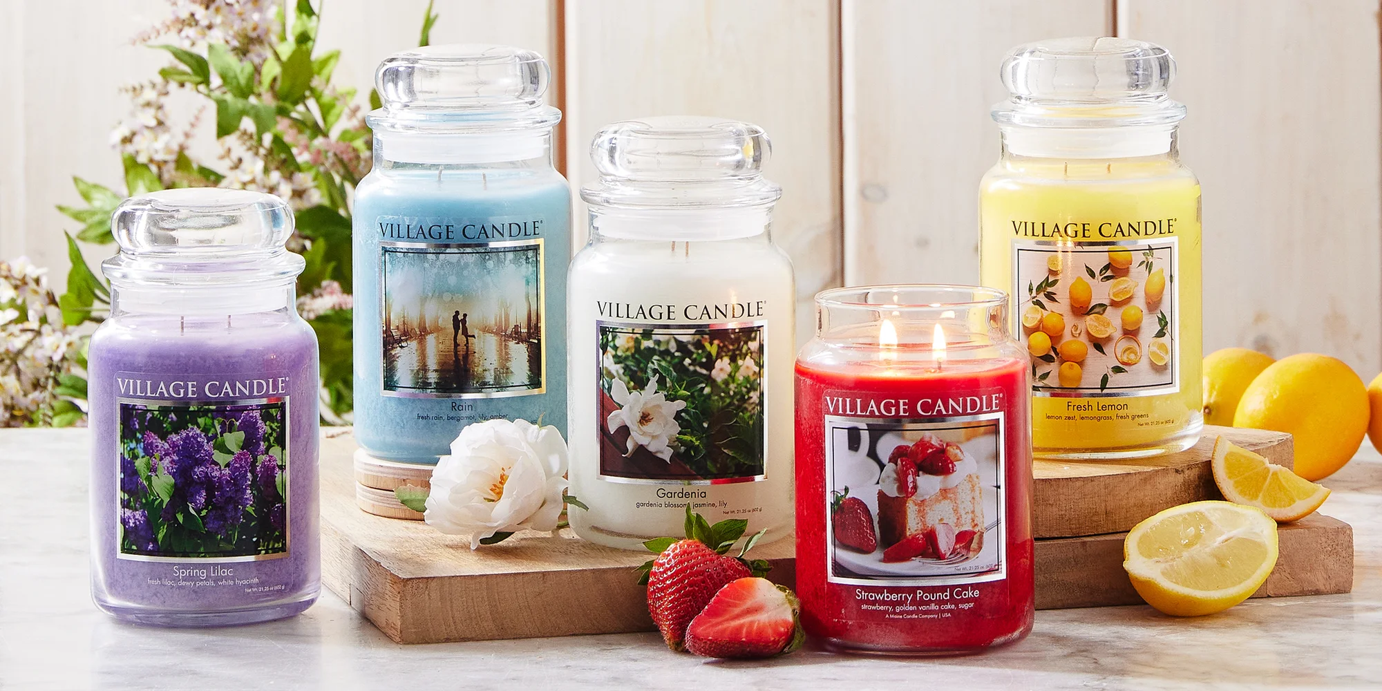 Five Village Candle jars on a wooden table: Spring Lilac, Rain, Gardenia, Strawberry Pound Cake and Fresh Lemon, with flowers, strawberries and lemons around them